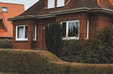 A cozy red brick house with a sloped roof and trimmed hedges in a suburban neighborhood.